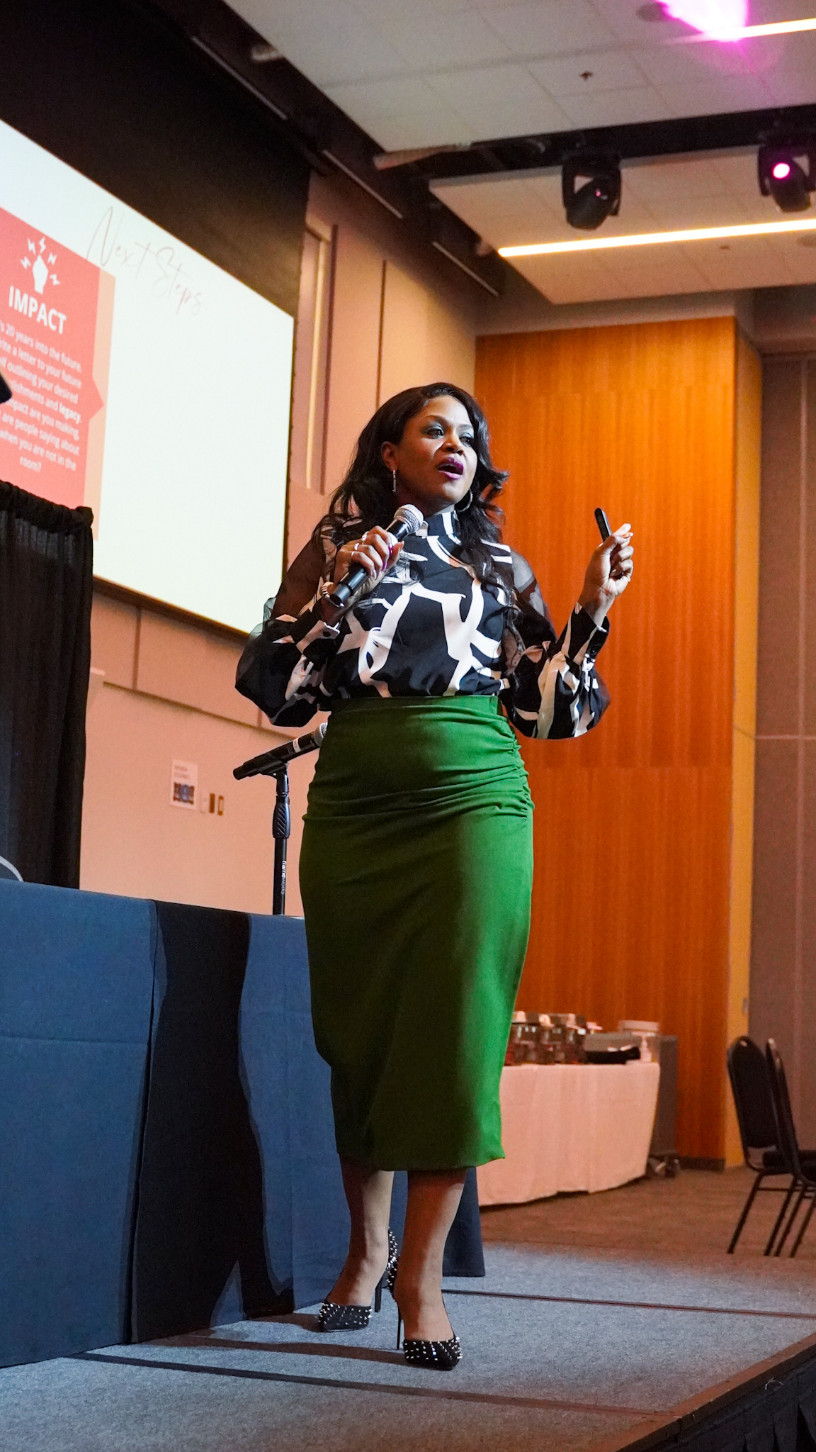 Monique Bryan speaking on stage with a microphone, IMPACT framework slide behind her